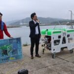 Kyushu Institute of Technology Center for Social Implementation of Future Robots head Yuya Nishida, center, explains the new Mega Bee submersible robot before its launch ceremony, at Waita fishing port in the Anya area of Wakamatsu Ward, Kitakyushu, Fukuoka Prefecture, April 10, 2026. (Mainichi/Yumiko Tani)