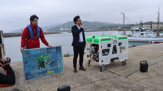 Kyushu Institute of Technology Center for Social Implementation of Future Robots head Yuya Nishida, center, explains the new Mega Bee submersible robot before its launch ceremony, at Waita fishing port in the Anya area of Wakamatsu Ward, Kitakyushu, Fukuoka Prefecture, April 10, 2026. (Mainichi/Yumiko Tani)