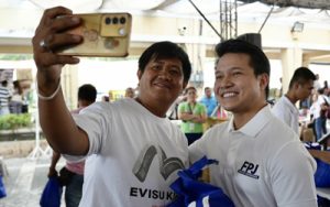 Two men take a selfie at an outdoor event; the man on the left holds a gold smartphone while both smile, with bystanders in the background.