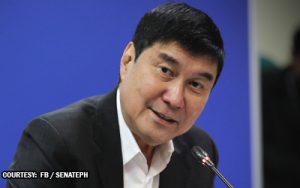 Man in a dark suit speaking into a microphone at a press conference, blue background behind him.