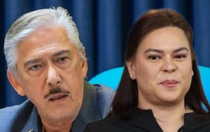 Two adults seated side by side at a formal event, the man with gray hair and mustache looking serious while the woman beside him smiles slightly. Backdrop is blue curtains.