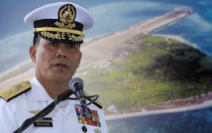 High-ranking naval officer in dress whites speaking into a microphone, with medals on his chest and an island airstrip backdrop.