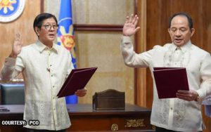 Two men in formal attire take an oath, raising their right hands while holding oath papers in a ceremonial setting with a flag in the background.