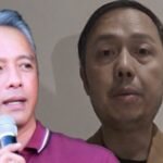 Two men on stage; left man with gray hair speaks into a microphone, wearing a maroon polo, while a second man stands beside him.