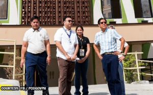 Four adults posing for a photo outside a green-and-white building, wearing badges and lanyards.