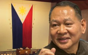 Close-up of a smiling man in a conference room, with the Philippine flag on the wall behind him.