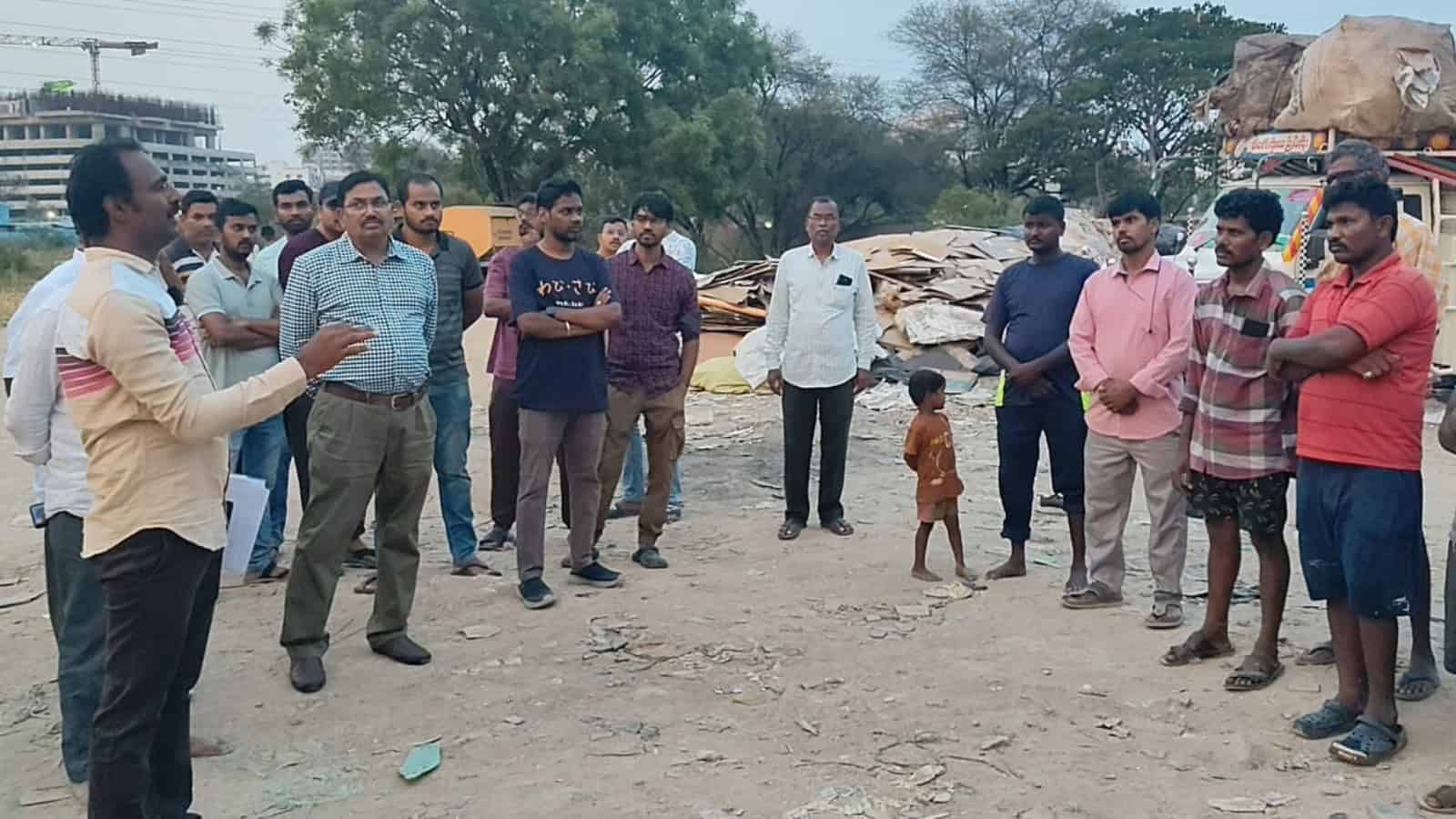 A detailed group of people gathered outdoors, listening to a speaker in a dusty area with scattered debris.