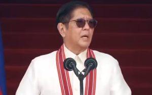 Man in a white suit with red trim and sunglasses speaks into two microphones on a stage.